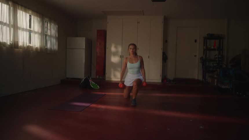 A woman in a workout tank top and shorts performs a lunge while holding dumbbells in a sunlit room