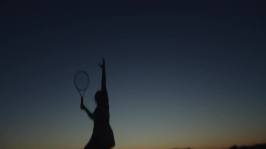 A female tennis player in silhouette hits a serve
