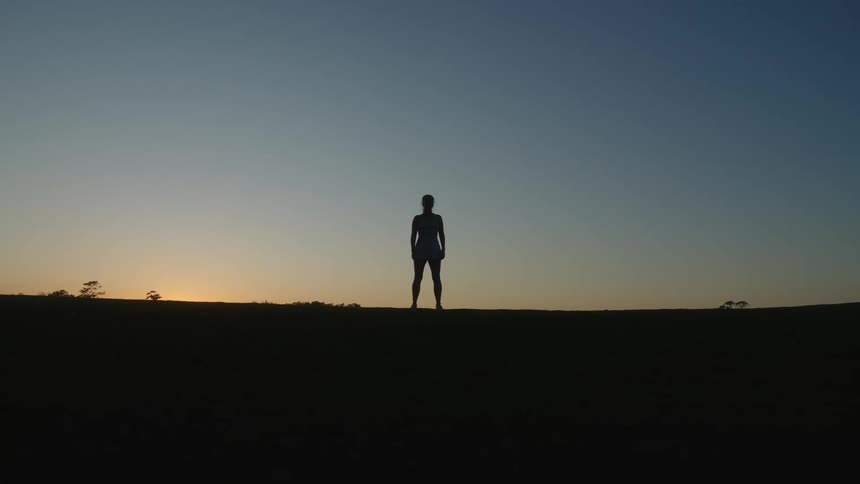 Silhouette of a woman standing on top of a hill against a dawn sky