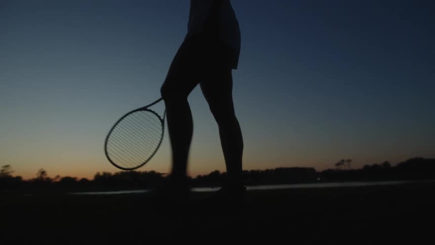 Female tennis player holding a tennis racket walks while silhouetted against the sky at night
