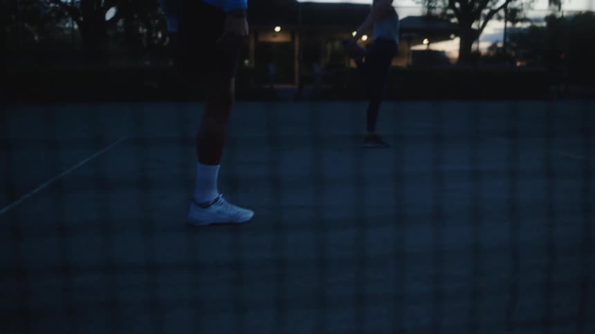 Two tennis players stretch on an outdoor court at dusk