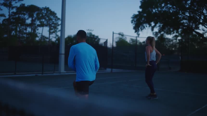 Man and woman stretch while on tennis court after dusk