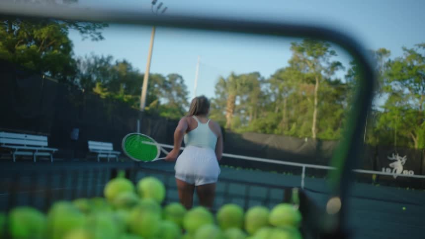 A tennis player serving a ball in the court at morning practice in front of a basket of tennis balls