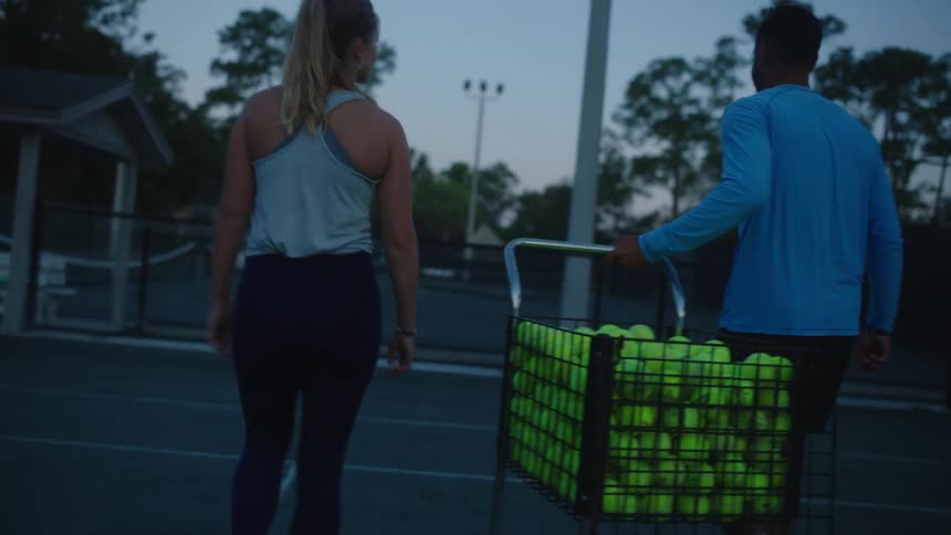 Two people preparing for a twilight tennis session with a man pushing a cart full of tennis balls