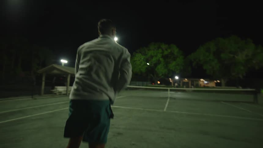 A man is playing tennis and hitting the ball with the racket at night