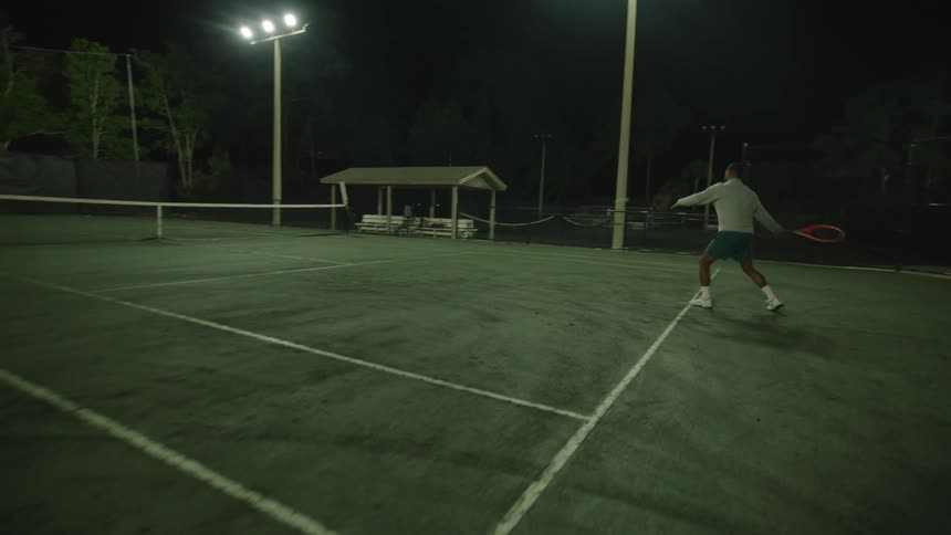 A man is playing tennis on a court at night and hitting the ball using his racket