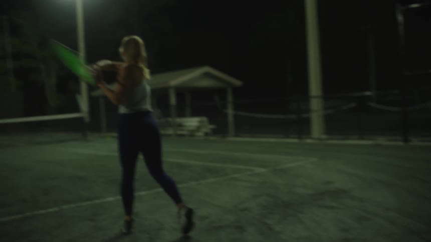 A woman swings a tennis racket playing on a welllit court at night