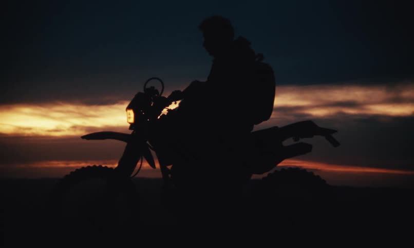 Silhouette of a man taking off his helmet while sitting on a motorbike against a red sky
