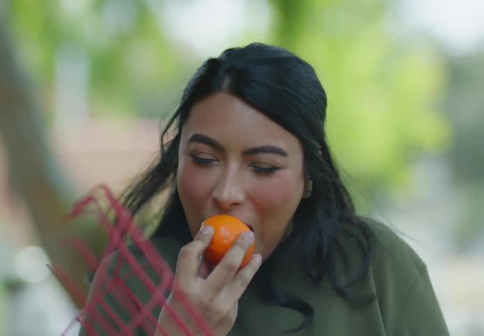 A woman with straight hair winces playfully as she takes a bite of a sour mandarin in the sunshine drenched garden