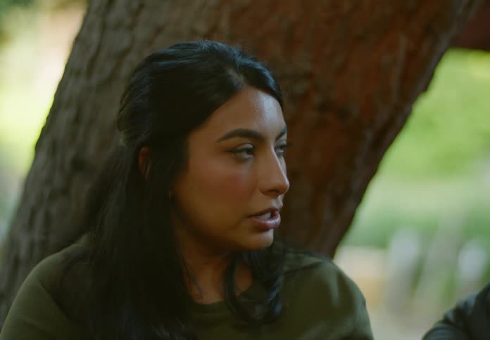 A woman with straight hair and a green shirt laughs while talking to a man in front of a tree