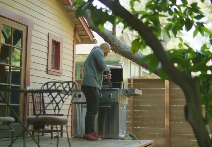 A bald man cleans a metal grill on a deck behind a house