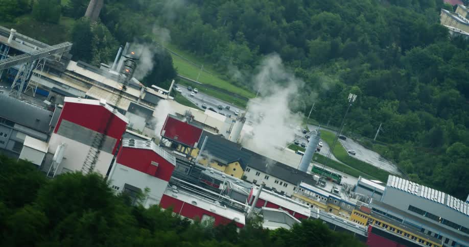 Some industrial buildings with white smoke coming out of chimneys and behind them there is a forest of trees with green leaves