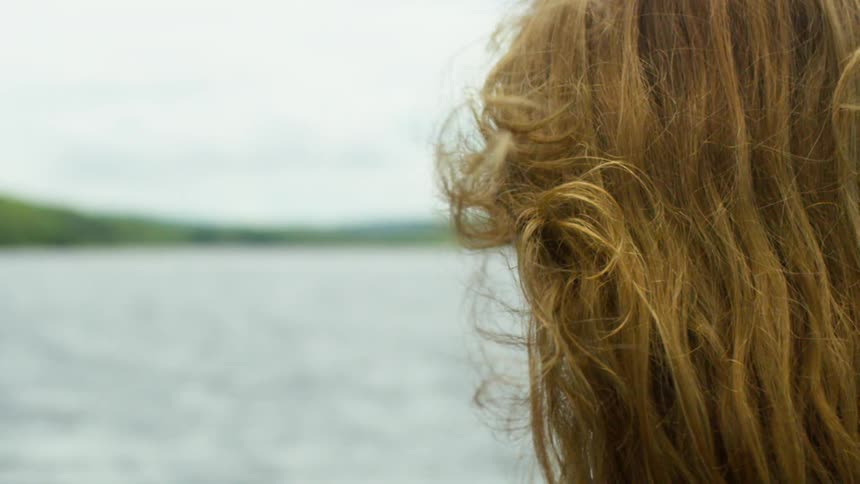 Redhead stands looking toward distant forest across the water on an overcast day