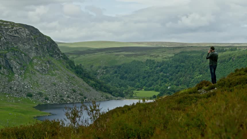 A man standing on a grassy hill overlooking a lake under a cloudy sky