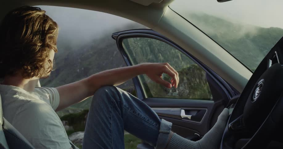 A man is sitting in a stopped car with the door open and he has his foot on the dashboard