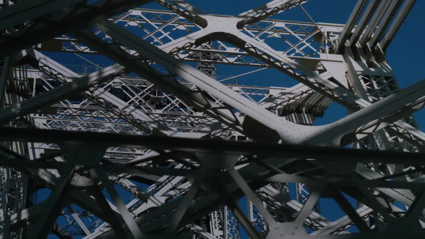 A metal bridge with a truss design stretches across a calm river under a clear blue sky