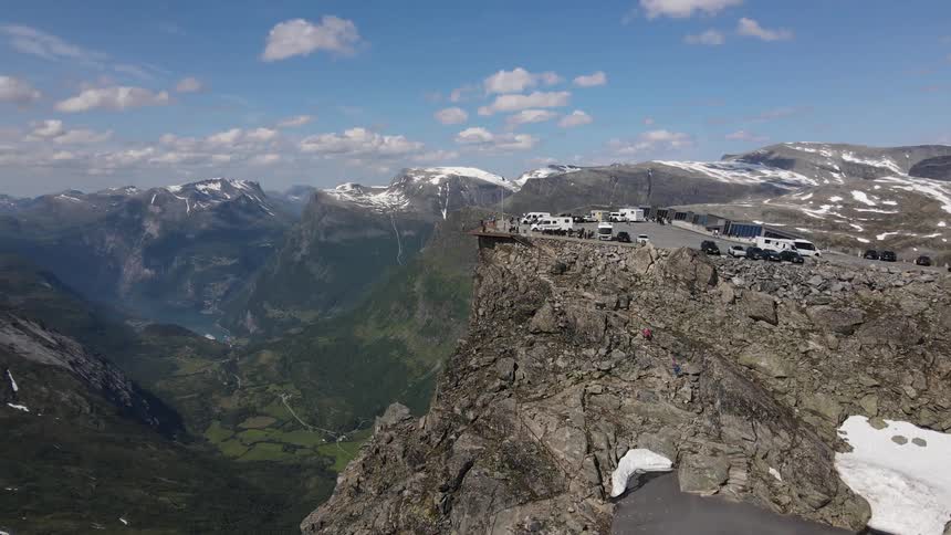 Aerial panorama of a rocky peak with parked cars and a panoramic view of several other snowy mountains during the day
