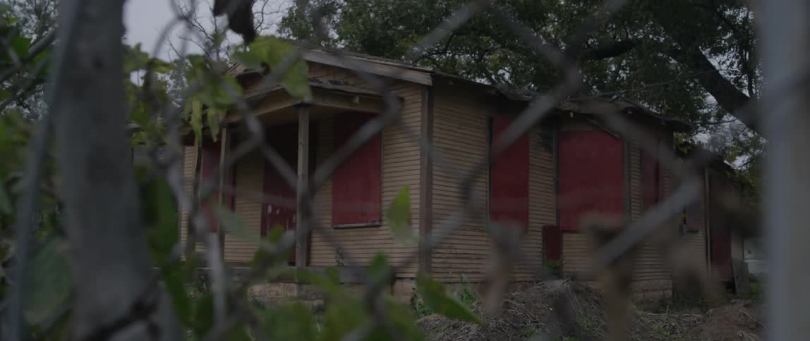 Abandoned home with red shutters seen through a wire fence
