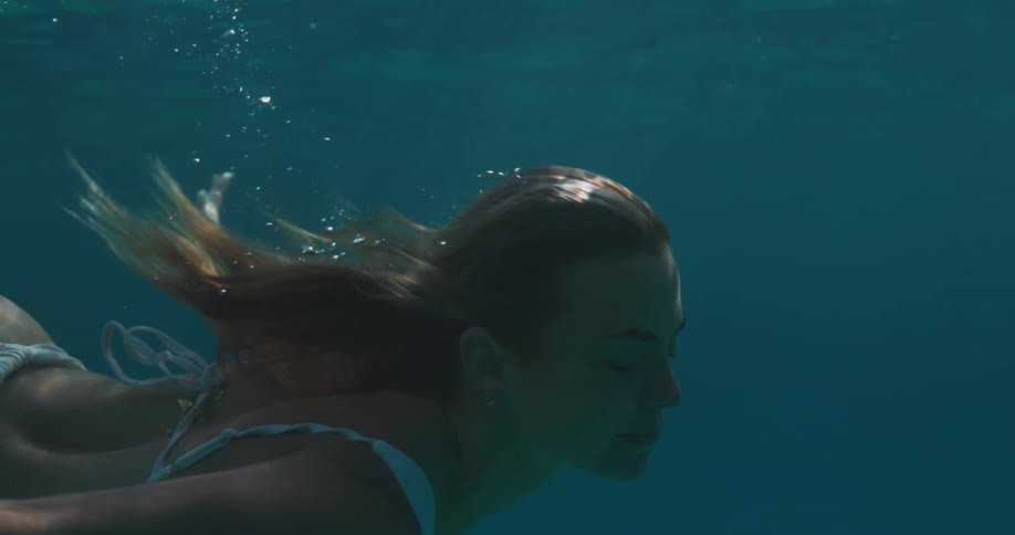 A young woman with her eyes closed is diving under the clear water with a few beams of sunlight hitting her