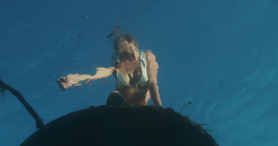 Upward view from inside the ocean of a woman in a bikini at the surface running her hand through the ocean waters