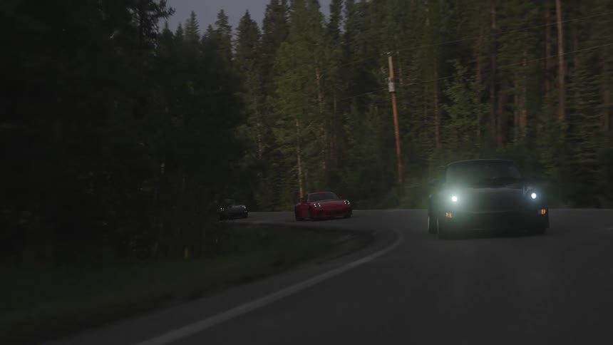 A black sports car driving on an asphalt street between pine forests and there are some other sports cars behind it