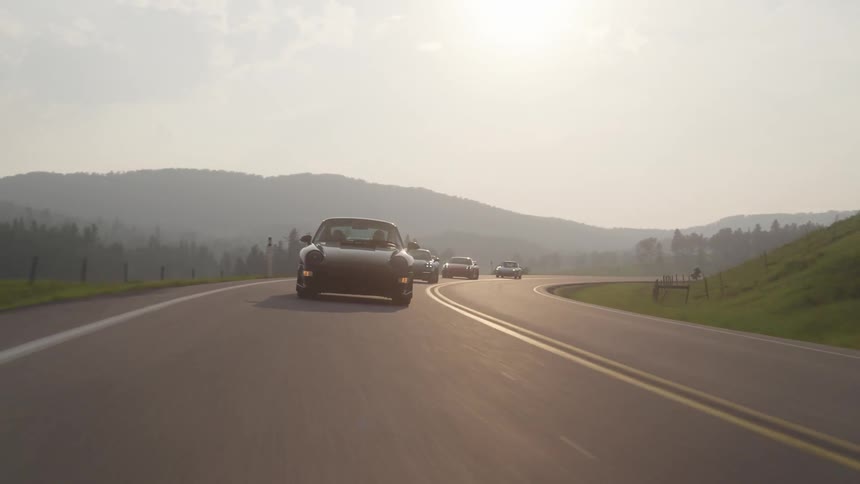 Porsches traveling along a paved road with mountains around at sunset