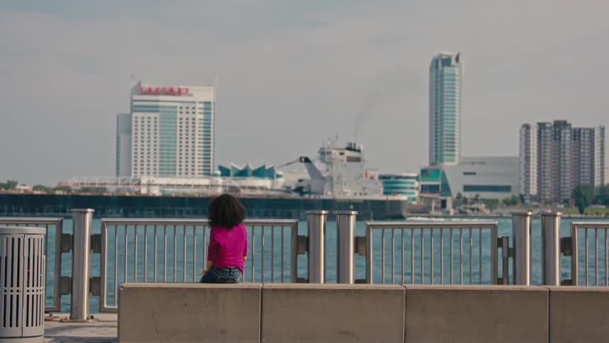 A woman in a pink shirt sits alone on a bench gazing across the water at a cityscape dominated by tall buildings and a large ship