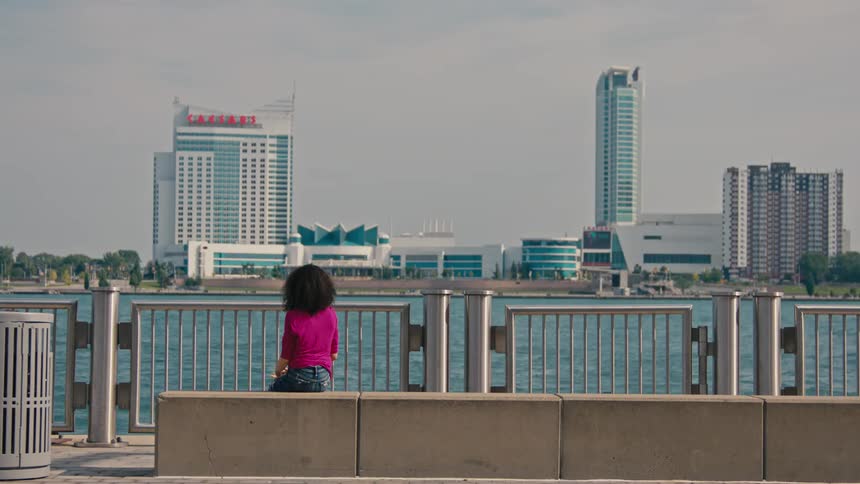 Woman sitting in a public environment by the sea with a jet ski passing by and skyscrapers of a metropolis on the horizon