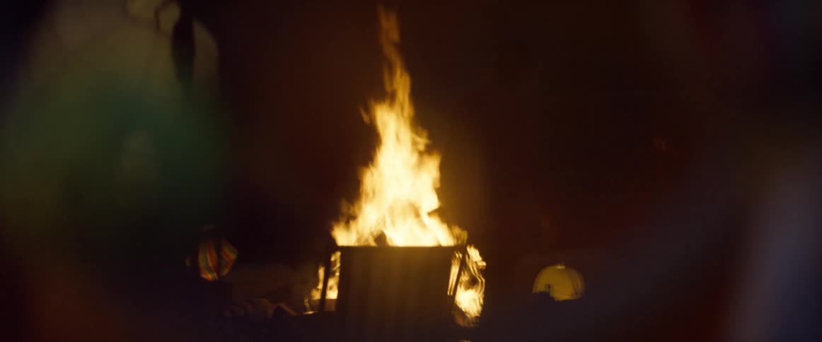 A man holding a bottle gets up from a lawn chair in front of a bonfire
