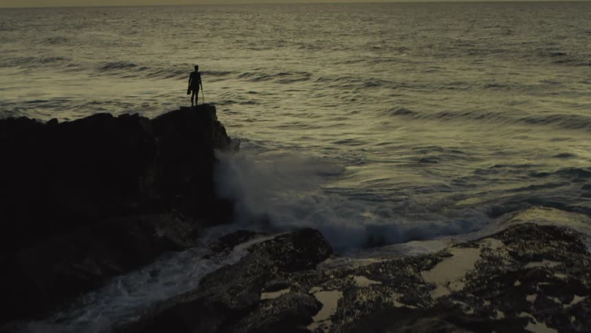 A man with a surfboard stares at the ocean from a rocky coastal outcropping