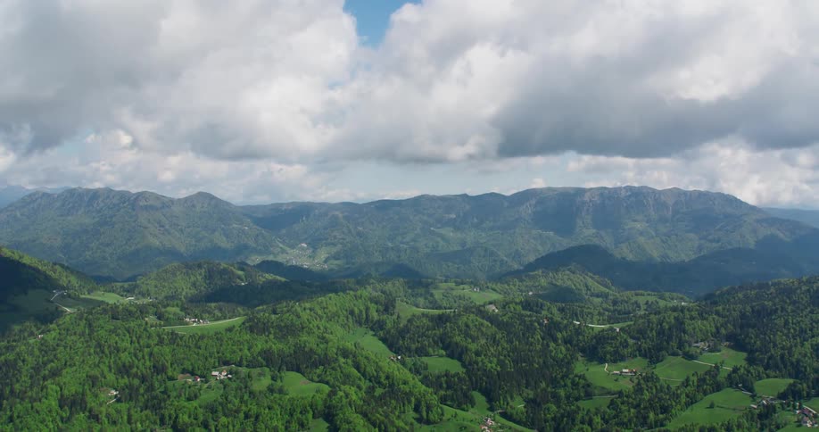 An aerial view of a region with hills and a mountain range with lots of vegetation and white clouds in the sky
