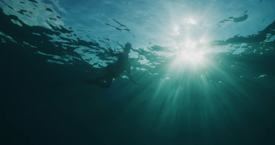 A person swimming in blue water seen from underwater with strong sunlight coming from the sky