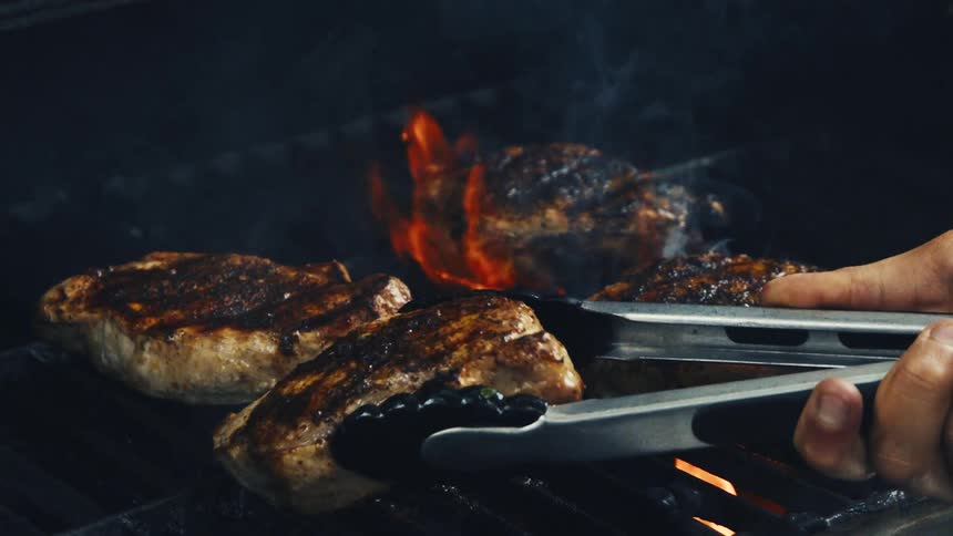 A persons hands flip steaks on the grill in slow motion