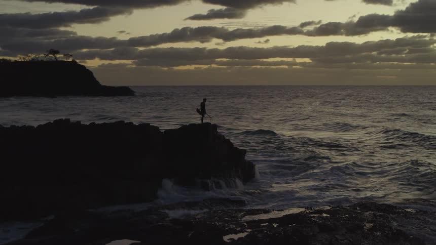 A lone fisherman silhouetted against the colorful sunset casts his line off a rocky cliff