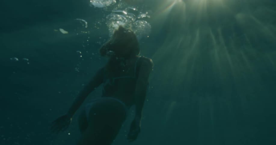 Woman in white bikini submerged in ocean waters heading towards the surface with oxygen bubbles around her