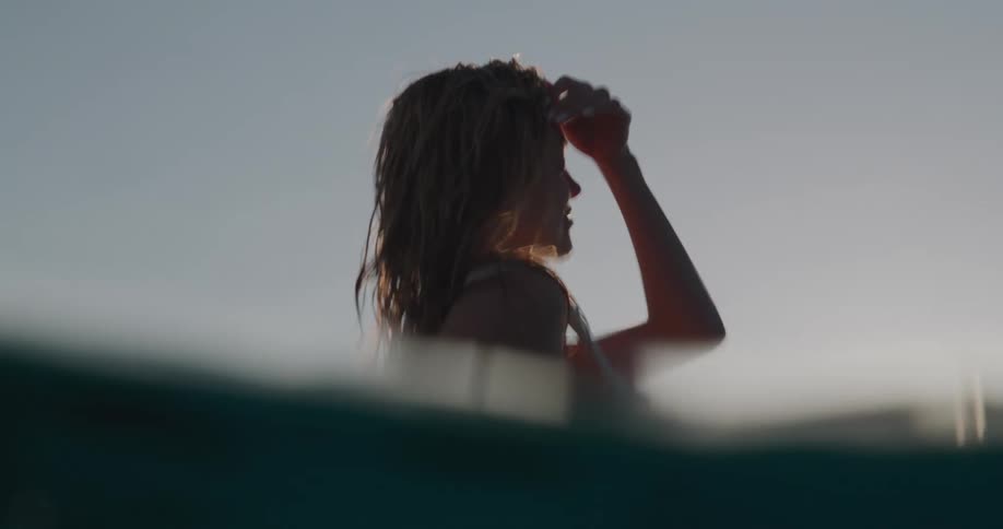 A woman is sitting and looking forward with her hand protecting her face from the sun and in front there are ripples of water