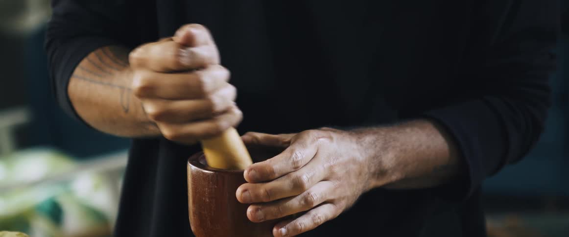 A man wearing a black sweater uses a large wooden mortar and pestle to grind ingredients