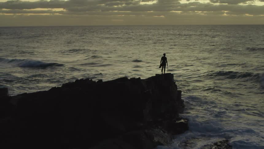 A man holding a surfboard stands on a coastal rocky outcropping