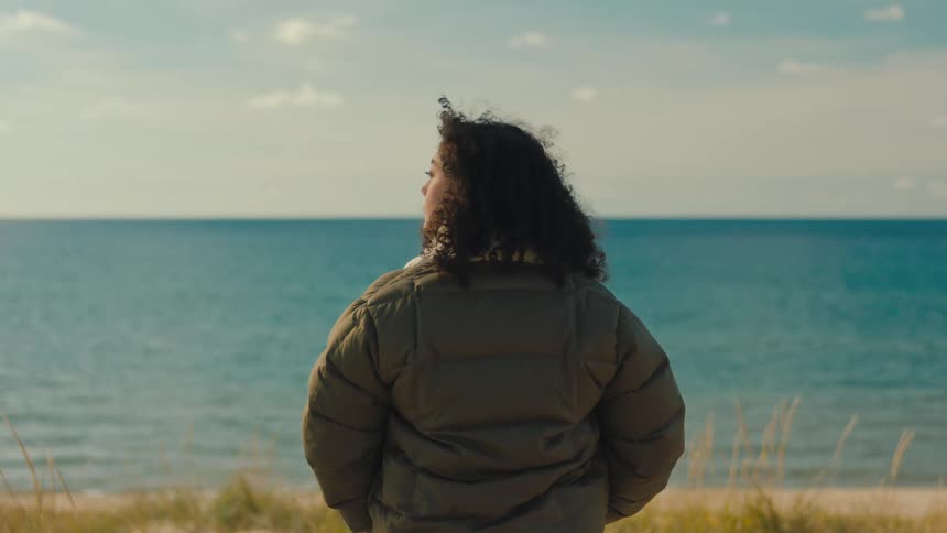 A woman with curly hair and a dark green coat watches the blue sea and sandy beach