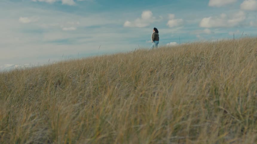 A woman wearing a white scarf a green jacket and jeans walks down a hill slope with brown grass