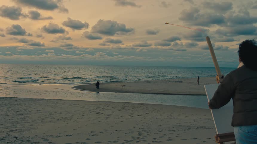 A painter on a beach preparing to paint a scene with two people flying a kite