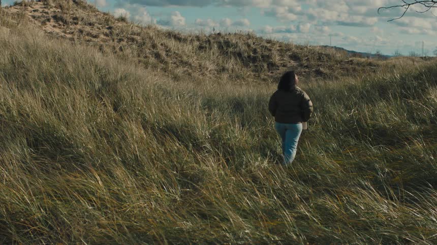 A woman with curly hair wearing a green coat and jeans walks down a grassy hill slope