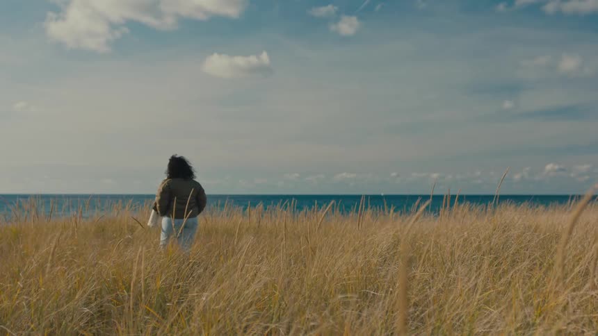 A woman with curly dark hair is walking through a field of tall grass under a blue sky with scattered clouds next to the ocean