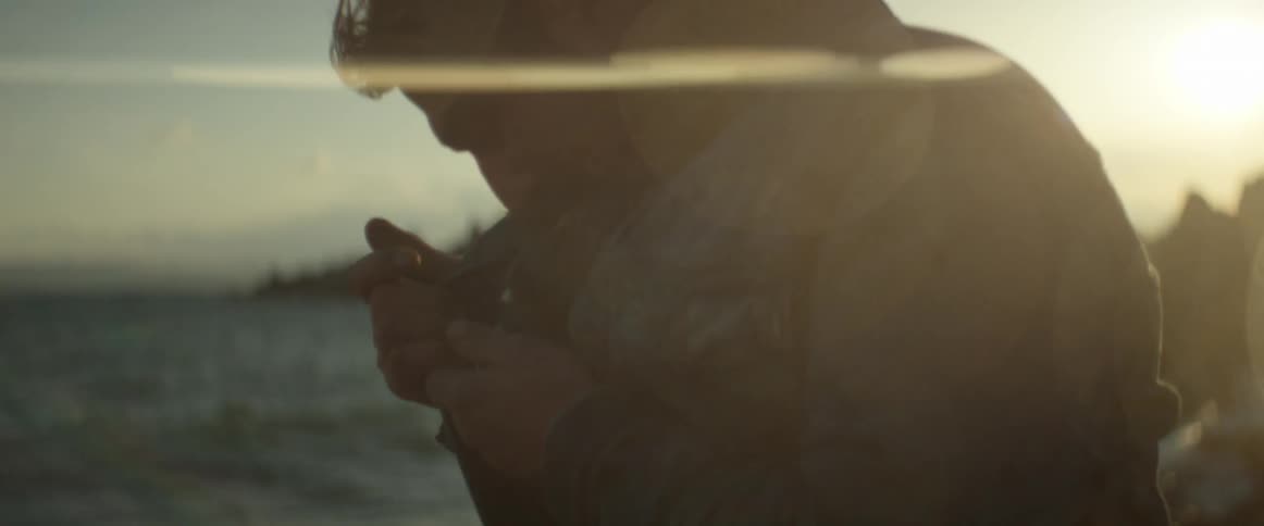 A man holding his coat out to shield the flame of his lighter as he lights a cigarette on a windy shoreline