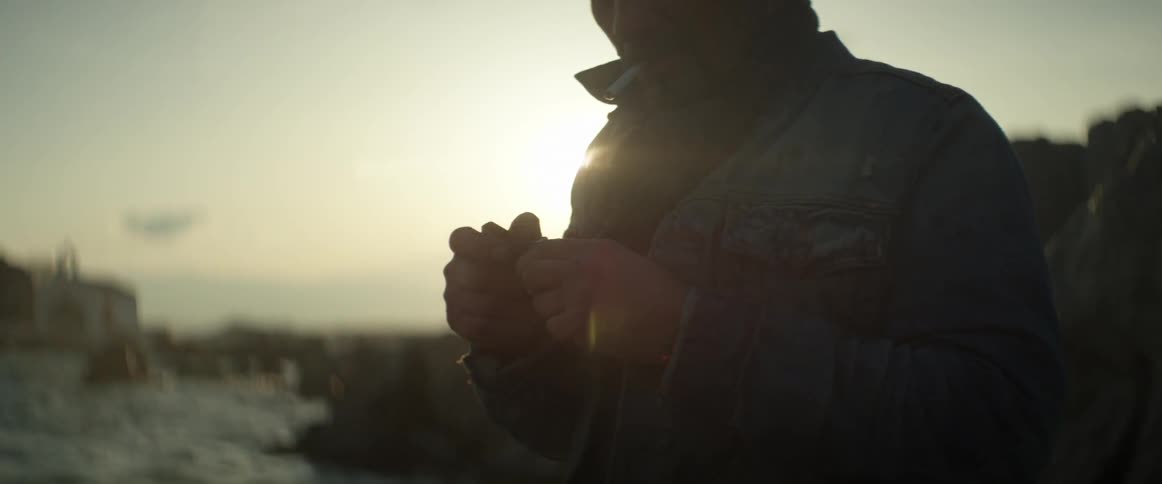 A man holding out his coat to protect his lighter's flame as he bends over to light his cigarette