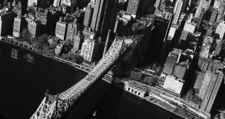 An aerial view of a city with the names of the buildings and a large suspension bridge over the water and everything is in black and white tones
