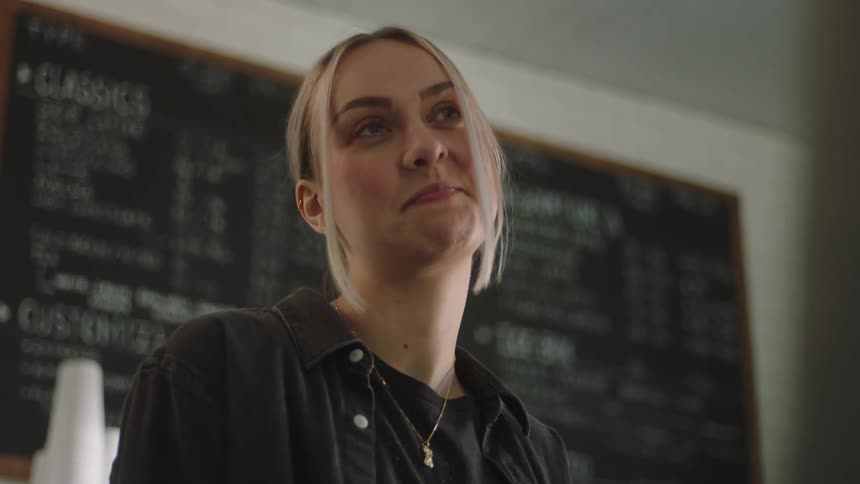 A blonde woman places a cup of coffee on the counter and begins to nod her head in confirmation