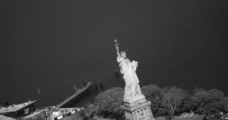 An aerial view of the statue of liberty which is on a small island in the sea and in black and white tones