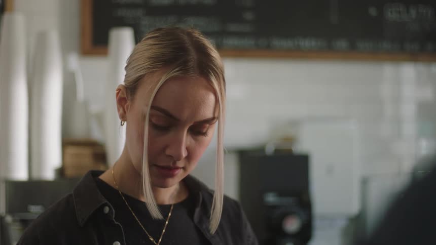 A blonde cashier looking down writing down something and then talking to someone in front of her with a smile on her face