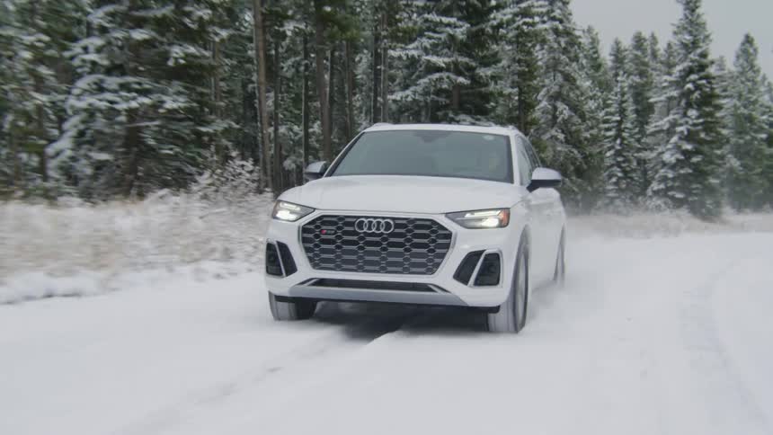 A white car driving on a street covered in snow and with pine trees on the left side
